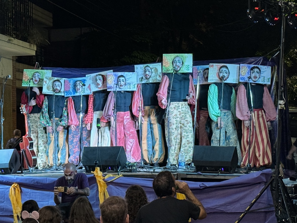 Murga performers on stage wearing vibrant costumes and currency face cutouts that show white painted faces.